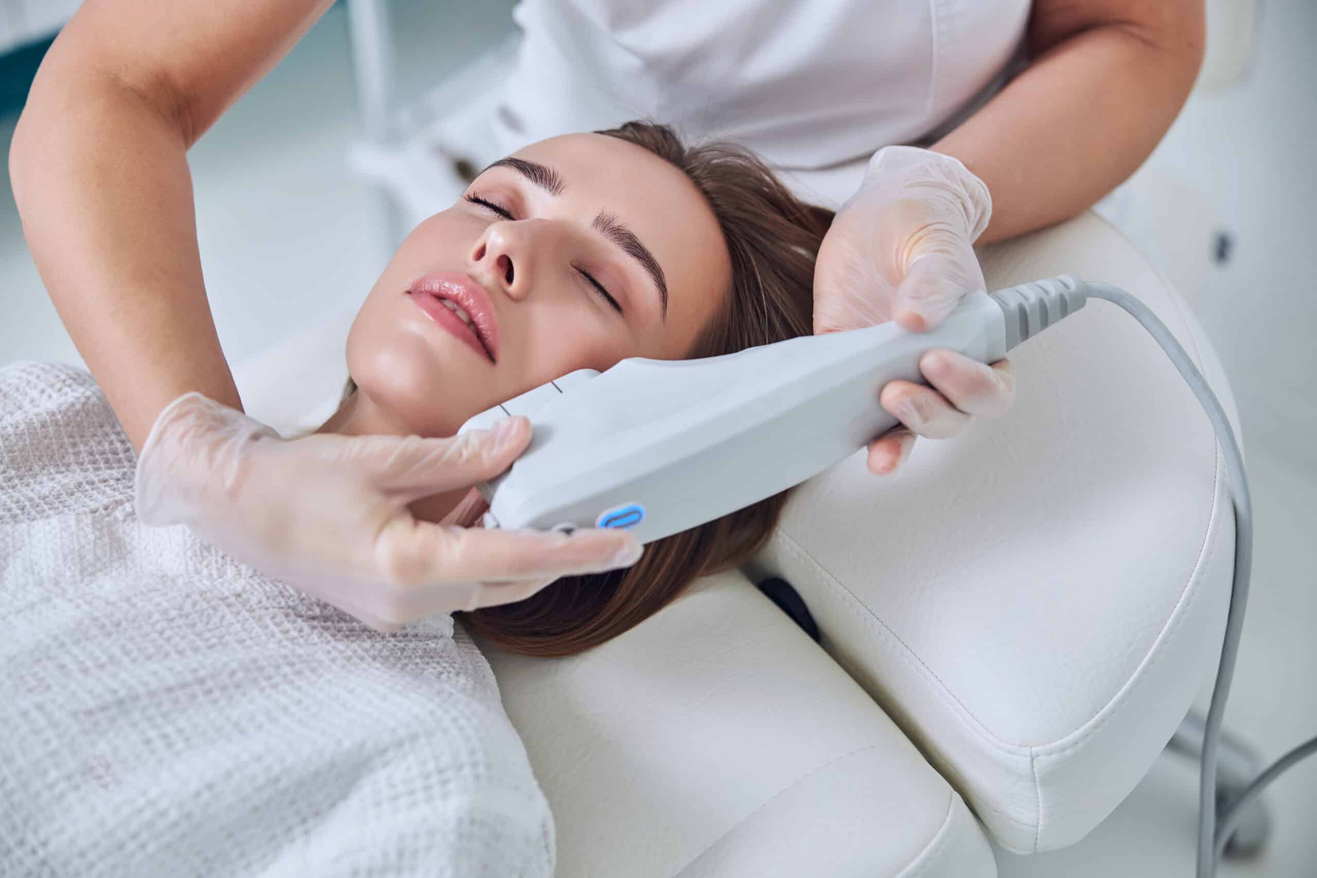 Close up top view portrait of elegant young female with close eyes lying on medicine chair while unrecognized beautician making spa treatment in hardware cosmetology